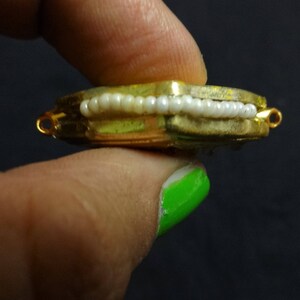 May include: A gold-toned, rectangular jewelry component with a row of white beads. The piece has small loops on each end and is held between fingers against a dark background. The jewelry component is likely for a necklace or bracelet.