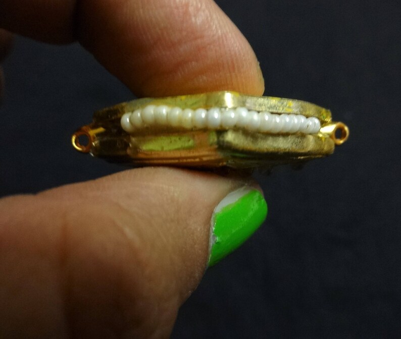 May include: A gold-toned, rectangular jewelry component with a row of white beads. The piece has small loops on each end and is held between fingers against a dark background. The jewelry component is likely for a necklace or bracelet.