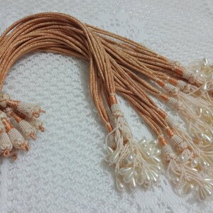 Peut inclure: Un lot de 20 cordons de couleur p&ecirc;che avec des perles blanches nacr&eacute;es et des glands. Les cordons mesurent environ 45 cm de long.