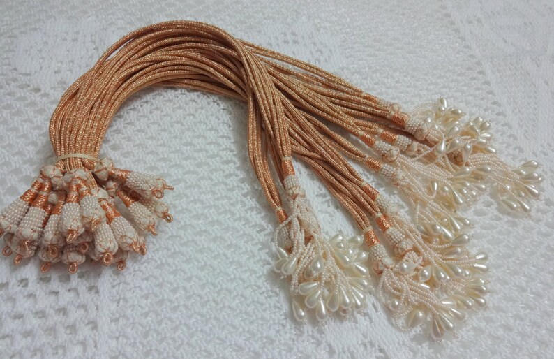 Peut inclure: Un lot de 20 cordons de couleur p&ecirc;che avec des perles blanches nacr&eacute;es et des glands. Les cordons mesurent environ 45 cm de long.