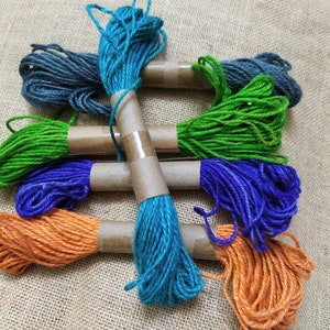 May include: A collection of six spools of twine in various colors, including teal, green, blue, orange, and gray. The twine is wrapped around cardboard spools and tied together with a brown string.