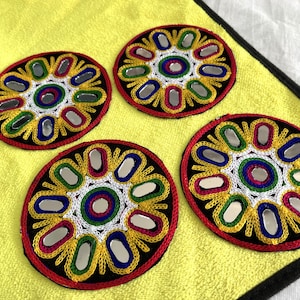 Puede incluir: Cuatro parches redondos, decorativos, bordados con centros de espejo. Los parches son negros con bordados amarillos, rojos, blancos y azules. Los centros de espejo están rodeados por un anillo de bordado blanco. Los parches están sobre un fondo amarillo.