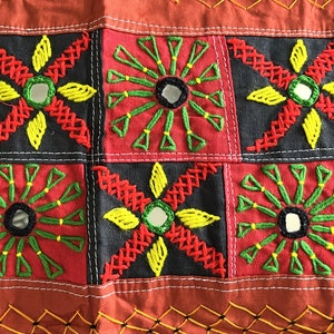 May include: A close-up of a colorful fabric with a patchwork design. The fabric is made of different colored squares, each with a different embroidered design. The designs include flowers, stars, and geometric shapes. The fabric is a vibrant mix of red, green, yellow, and black.