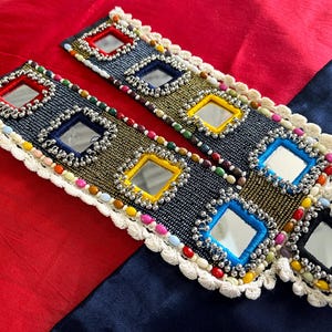 Può includere: Due bordure di perline con un bordo in pizzo bianco. Ogni bordura presenta una fila di piccoli specchi quadrati circondati da perline colorate e dettagli argentati.