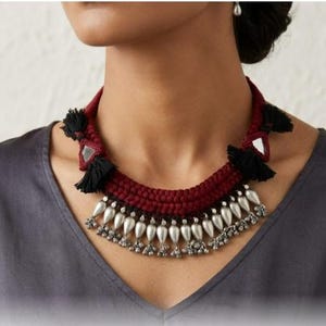 Op de afbeelding: Handgemaakte ketting met een gevlochten bordeauxrode koord, zwarte kwastjes en zilverkleurige druppelvormige hangers. De ketting heeft een uniek ontwerp met spiegelende elementen.