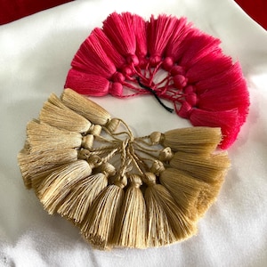 Puede incluir: Dos borlas decorativas, una rosa brillante y otra beige, dispuestas sobre una superficie blanca. Cada borla está hecha de múltiples hebras de hilo, atadas juntas en la parte superior. La borla rosa está extendida, mientras que la borla beige tiene una forma similar.