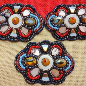 May include: Three beaded flower-shaped embellishments with a central white circle surrounded by colorful beads, shells, and stones. The embellishments are on a brown burlap background.