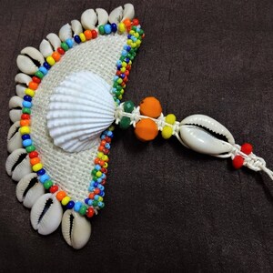 May include: A handmade fan-shaped ornament made of cowrie shells, colorful beads, and a white seashell. The ornament is attached to a white cord with a knot at the end.