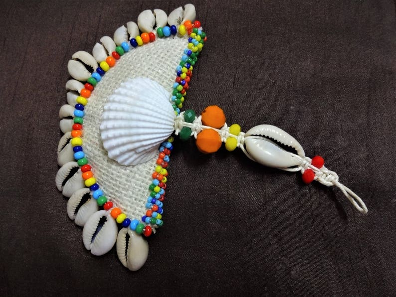 May include: A handmade fan-shaped ornament made of cowrie shells, colorful beads, and a white seashell. The ornament is attached to a white cord with a knot at the end.