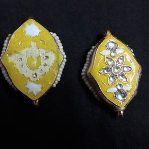 May include: Two yellow and gold-toned earrings with intricate designs. One earring features a white design, while the other showcases a floral pattern with clear gemstones. Both are outlined with small, white beads.