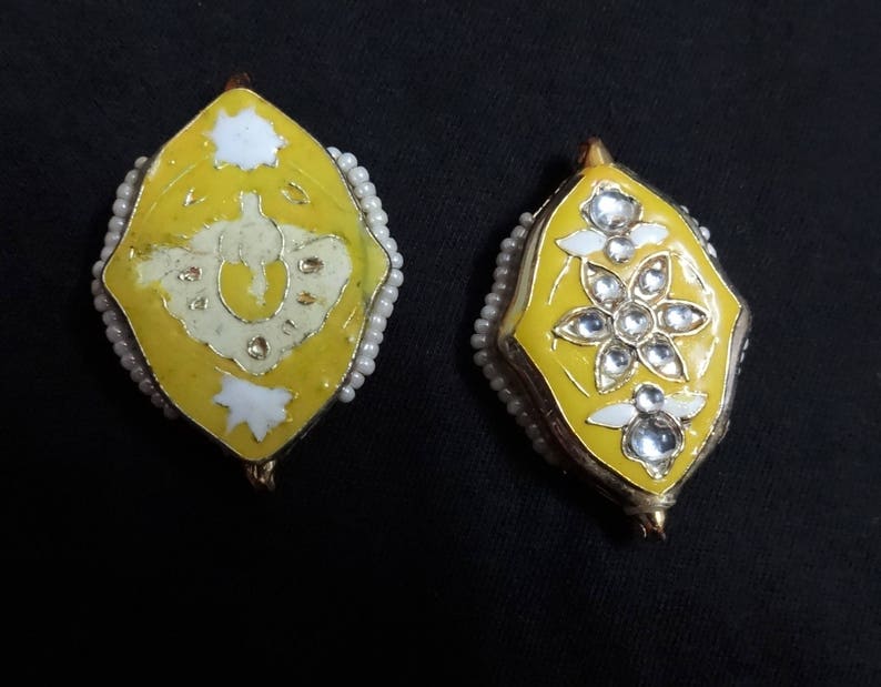 May include: Two yellow and gold-toned earrings with intricate designs. One earring features a white design, while the other showcases a floral pattern with clear gemstones. Both are outlined with small, white beads.