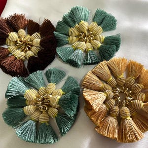 Könnte beinhalten: Vier dekorative Quastenornamente in Braun-, Grün- und Goldtönen. Jedes Ornament hat eine zentrale Ansammlung goldfarbener Perlen, die von strahlenförmigen Quasten umgeben sind. Die Ornamente sind auf einer weißen Oberfläche angeordnet.