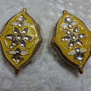 May include: Pair of ornate yellow earrings with a floral design. Each earring features a yellow enamel base, a gold-colored frame, and is adorned with clear, sparkling gemstones and white accents. The edges are lined with small, white beads.