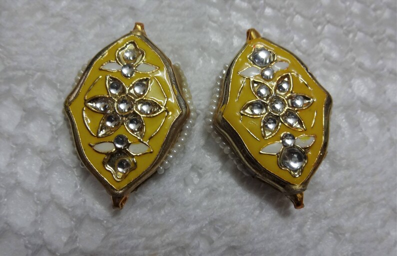 May include: Pair of ornate yellow earrings with a floral design. Each earring features a yellow enamel base, a gold-colored frame, and is adorned with clear, sparkling gemstones and white accents. The edges are lined with small, white beads.