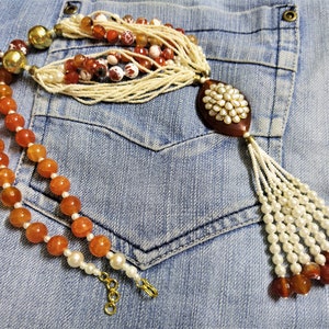 Op de afbeelding: Een meerstrengs ketting met een grote, ovale hanger. De hanger is gemaakt van bruine agaat en is bezet met witte parels. De ketting is gemaakt van witte parels, bruine agaat kralen en gouden kralen. De ketting ligt op een blauwe spijkerbroek.