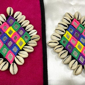 May include: Two decorative diamond-shaped ornaments. Each features a colorful, geometric pattern with small mirrors and is surrounded by seashells. One is on a magenta background, the other on white.