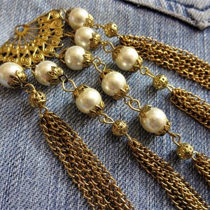 May include: A gold-toned metal pendant with a fan-shaped design and multiple chains. The pendant is adorned with white pearl-like beads.