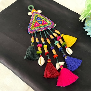 May include: A colorful, handcrafted textile ornament with a triangular top and dangling tassels. The top features embroidered patterns in pink, blue, yellow, and green. Tassels are in various colors, including red, pink, yellow, and blue, with wooden beads and seashells.