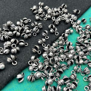 May include: A collection of small, silver-colored metal bead caps with a flower-like design. Each cap has a small loop for attaching to other components. The beads are scattered on a dark gray and teal fabric surface.