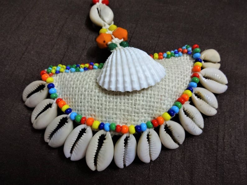 May include: A colorful beaded necklace with a white seashell pendant and a burlap backing. The beads are in shades of red, orange, yellow, green, blue, and purple.