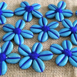 May include: A collection of blue and purple flower-shaped buttons with a textured surface. The buttons are arranged on a brown burlap background.
