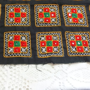 May include: Black fabric with a repeating pattern of embroidered squares. Each square features a red, green, and white design with small round holes.