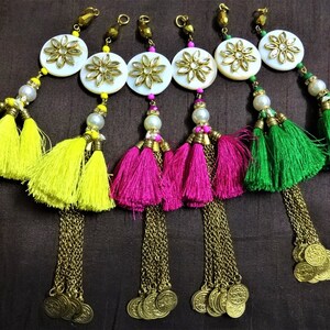 Op de afbeelding: Vijf decoratieve kwastjes met gouden kettingen en muntaccenten. Elk kwastje heeft een witte ronde kraal met een gouden bloemdessin en een parelkraal. De kwastjes zijn geel, roze, groen en twee geel.