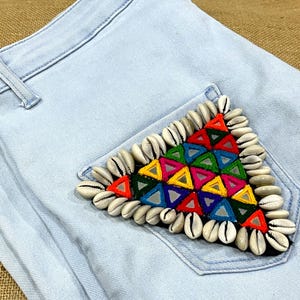 May include: Light blue denim jeans with a decorative pocket featuring a triangular design. The triangle is adorned with colorful geometric shapes and bordered by small white seashells. The jeans are made of a light wash denim.
