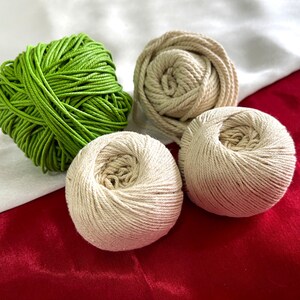 May include: Four balls of yarn in shades of green and beige rest on a red and white fabric background. The green yarn is a vibrant, tightly wound ball, while the beige yarn is in a looser, more natural form. Ideal for crafting projects.
