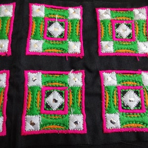以下が含まれることがあります： 緑、白、オレンジ、ピンクの糸で刺繍された正方形のパッチが5つ。パッチは黒い布地に縫い付けられています。
