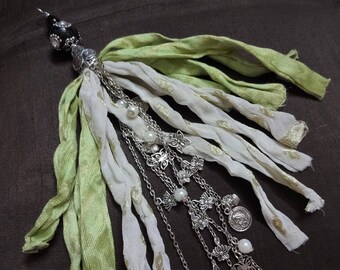 Puede incluir: Un borla de tela verde y blanca con cadenas y abalorios plateados, incluyendo mariposas y diseños florales. La borla está adornada con cuentas de perlas blancas.