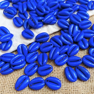 May include: A collection of vibrant blue cowrie shell beads. These oval-shaped beads have a glossy finish and a central slit, scattered on a white cloth and a textured, tan surface. Ideal for jewelry making or craft projects.