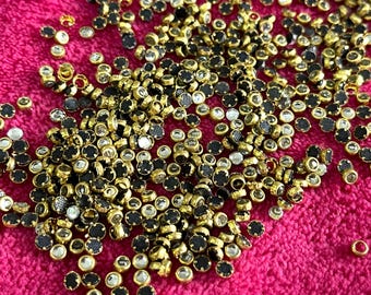 May include: A large pile of round, clear rhinestones with gold settings and black centers. The rhinestones are scattered on a pink surface.