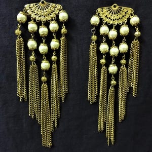 K&ouml;nnte beinhalten: Ein Paar goldfarbene Ohrringe mit wei&szlig;en Perlen und herabh&auml;ngenden Ketten. Die Ohrringe haben oben ein dekoratives F&auml;cherdesign.