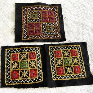 Può includere: Tre quadrati di tessuto nero con ricami intricati in filo rosso, verde e oro. I quadrati sono decorati con motivi geometrici e piccoli fori.