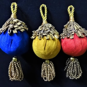 May include: Three decorative tassels in blue, yellow, and red. Each tassel has a round, fabric-covered ball with a gold-coloured fringe and a beaded tassel. The tassels are suspended by a gold rope loop.
