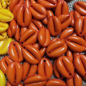 Puede incluir: Primer plano de un montón de conchas de cauri naranja y amarillo. Las conchas son lisas y brillantes, y tienen un agujero natural en la parte superior.