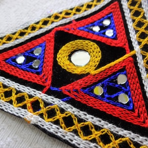 May include: A triangular fabric patch with intricate embroidery in red, blue, and yellow thread. The patch features a black background with small round mirrors sewn into the design.