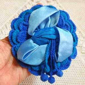 Puede incluir: Una flor de crochet azul con un lazo de cinta de satén azul. La flor tiene un diseño texturizado y con volantes y está adornada con pompones azules.