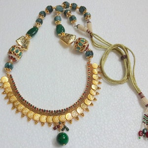 Peut inclure: Un collier en or avec un pendentif en perles vertes et rouges. Le collier pr&eacute;sente un motif de pi&egrave;ces d'or avec des pierres vertes et rouges.