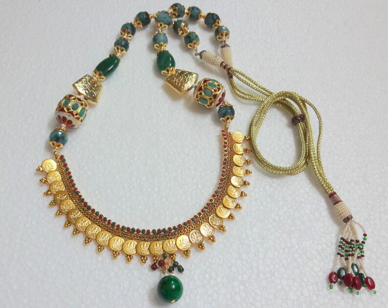 Peut inclure: Un collier en or avec un pendentif en perles vertes et rouges. Le collier pr&eacute;sente un motif de pi&egrave;ces d'or avec des pierres vertes et rouges.