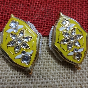 May include: A pair of yellow and gold earrings with a floral design. The earrings have white accents and clear, round gemstones. The edges are lined with small white beads. The earrings are set against a textured, neutral background.
