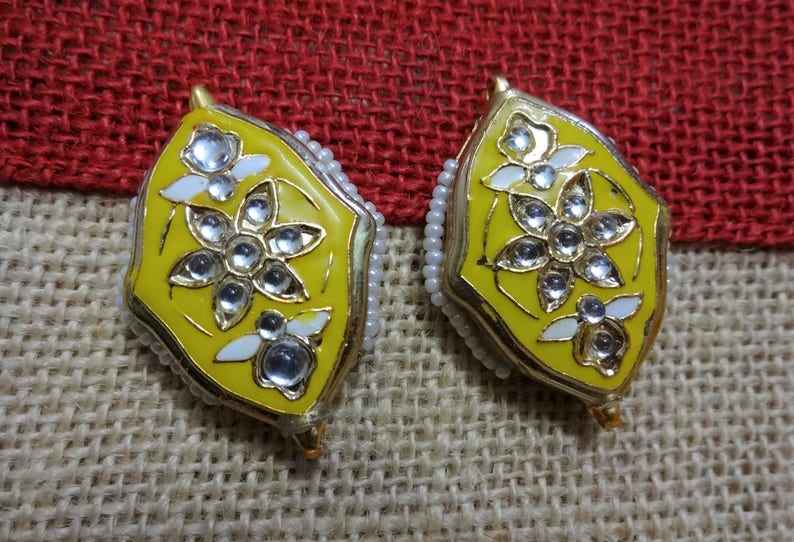 May include: A pair of yellow and gold earrings with a floral design. The earrings have white accents and clear, round gemstones. The edges are lined with small white beads. The earrings are set against a textured, neutral background.