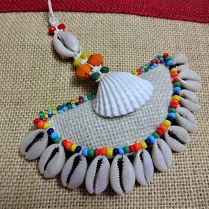 May include: A handmade necklace made of cowrie shells, colorful beads, and a white seashell. The necklace is displayed on a burlap background.