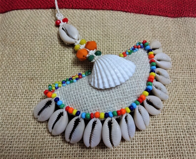 May include: A handmade necklace made of cowrie shells, colorful beads, and a white seashell. The necklace is displayed on a burlap background.