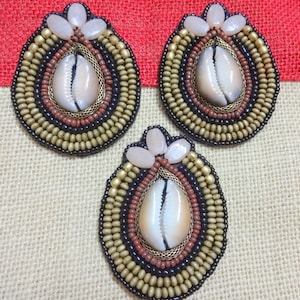 May include: Three handcrafted oval-shaped brooches. Each brooch features a central cowrie shell, surrounded by layers of beads in black, brown, gold, and beige. The top of each brooch has three light pink, petal-shaped accents.