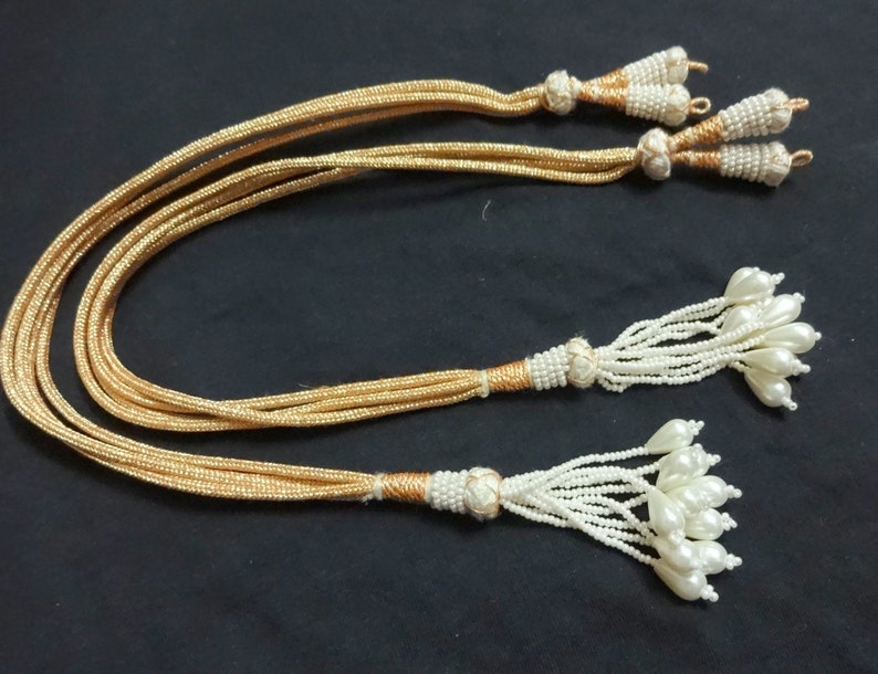 Peut inclure: Trois cordons dor&eacute;s avec des glands en perles blanches. Chaque gland est termin&eacute; par un groupe de perles nacr&eacute;es.