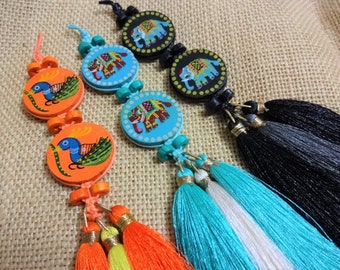 May include: Four colorful wooden ornaments with tassels. Each ornament features a different design: a peacock, an elephant, and two elephants. The ornaments are strung together with beads and have colorful tassels.