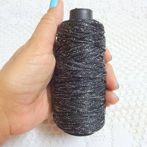 Puede incluir: Una bobina de hilo negro con purpurina plateada. El hilo est&aacute; enrollado firmemente alrededor de la bobina.