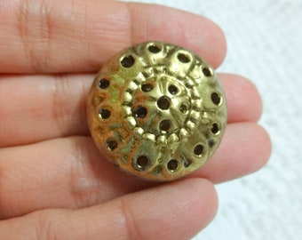 May include: A gold-colored metal button with a decorative, circular design featuring a raised, concentric pattern of circles and holes.